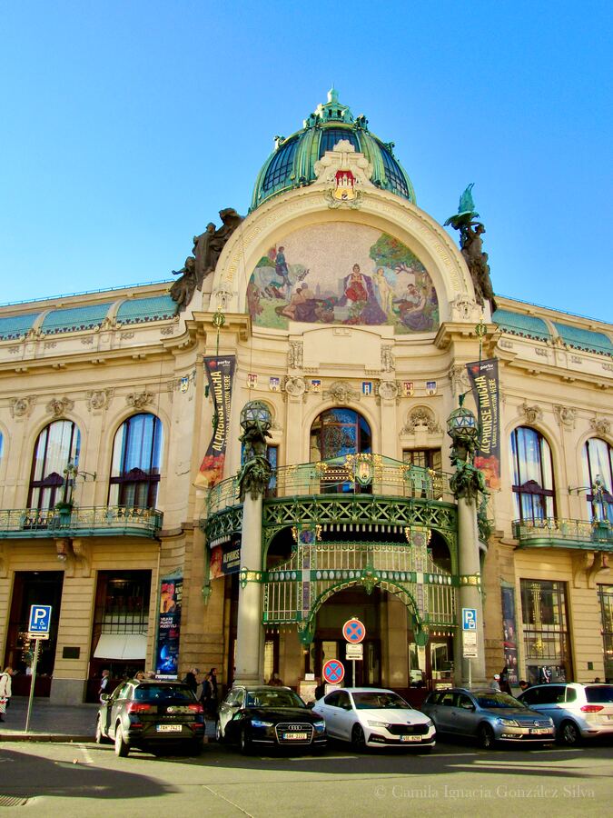Front view of the Municipal House in Prague with its dome, mosaic, ornate ironwork entrance, banners, and parked cars along the street