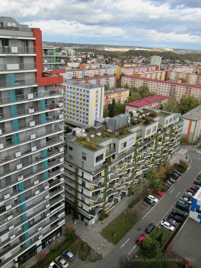 High-angle view of contemporary residential buildings in Prague with terraces, integrated vegetation, internal streets, parked cars, and older housing blocks in the background