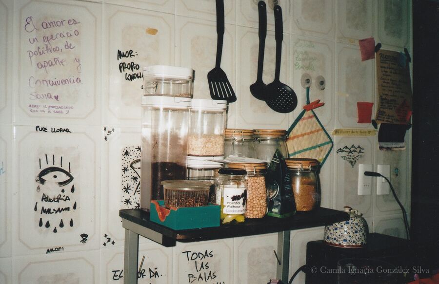 Kitchen corner in Ñuñoa with jars of stored food, hanging utensils, a kettle on the stove, and handwritten notes and drawings covering the wall