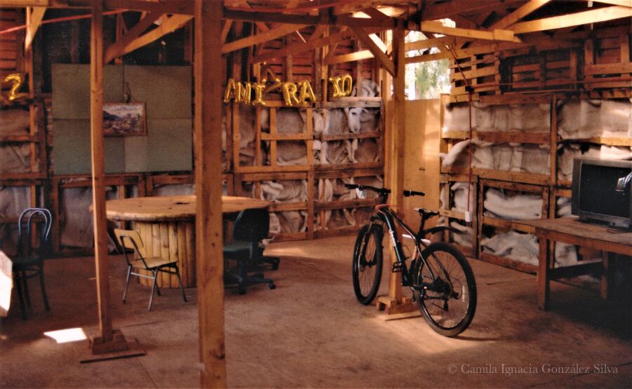 Interior of Cootramap’s 2022 self-built headquarters in La Pintana, with exposed wooden beams, a reused cable-spool table, mismatched chairs, a bicycle, and a television