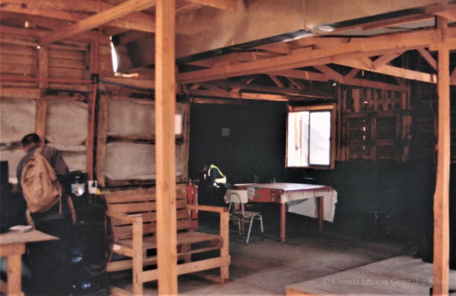 Interior of Cootramap’s 2022 self-built headquarters in La Pintana, with exposed wooden beams, unfinished insulated walls, a table, chairs, a wooden bench, and a worker with a backpack