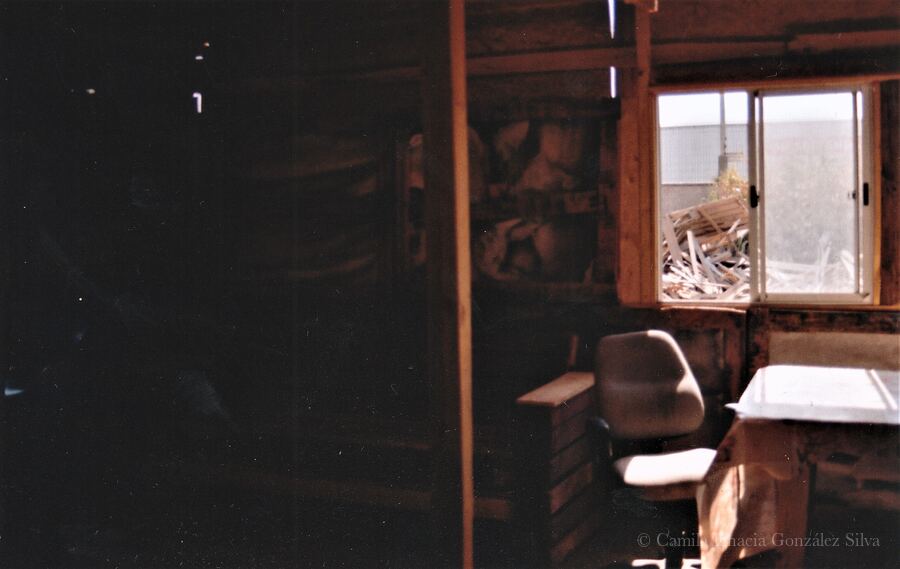 Interior of Cootramap’s 2022 self-built headquarters in La Pintana, with unfinished wooden walls, an office chair, a small drawer unit, a table, and an open window facing stacked materials outside
