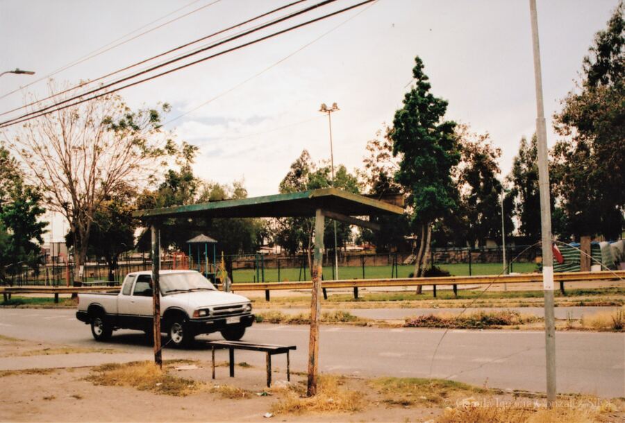 Street scene in Puente Alto with a roadside bus stop structure, a pickup truck, overhead wiring, and a fenced green area in the background