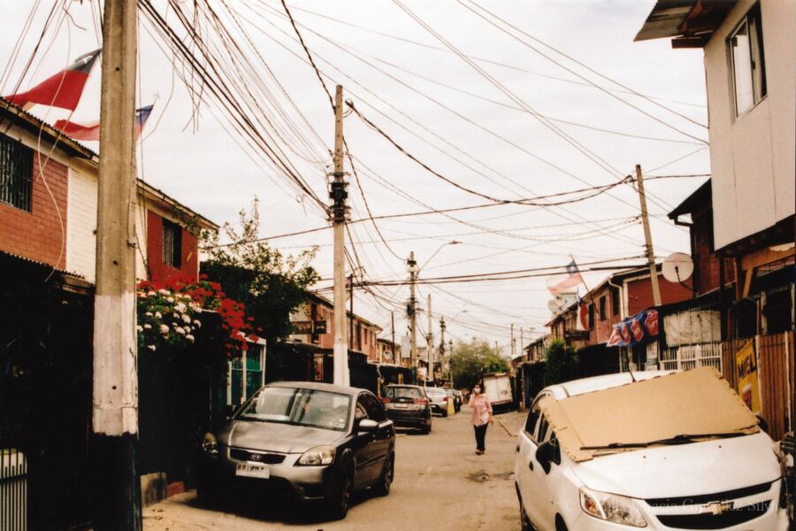 Residential street in Puente Alto with parked cars, attached houses, Chilean flags, and dense overhead wiring