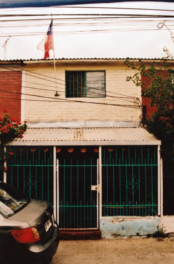 Two-story residential house in Puente Alto with metal fencing, overhead wiring, and a Chilean flag