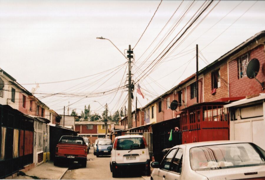 Residential street in Puente Alto with attached houses, parked cars, and dense overhead wiring