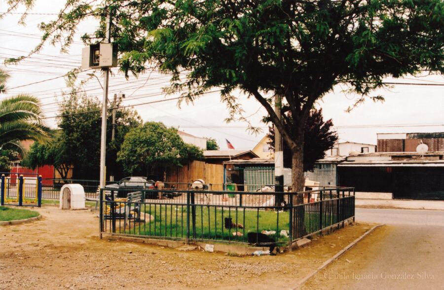 Small fenced green space in Puente Alto with residential housing, visible infrastructure, and a street shrine