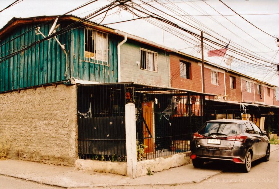 Row of attached houses in Puente Alto with metal fencing, dense overhead wiring, a parked car, and a Chilean flag