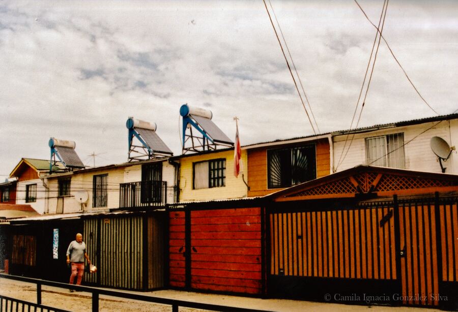 Row of attached houses in Puente Alto with metal enclosures, rooftop solar water heaters, overhead wiring, and a pedestrian