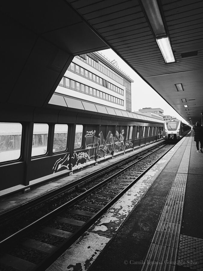 Covered train platform in Berlin with railway tracks, tactile paving, graffiti-covered barriers, office buildings, and a train approaching in the distance